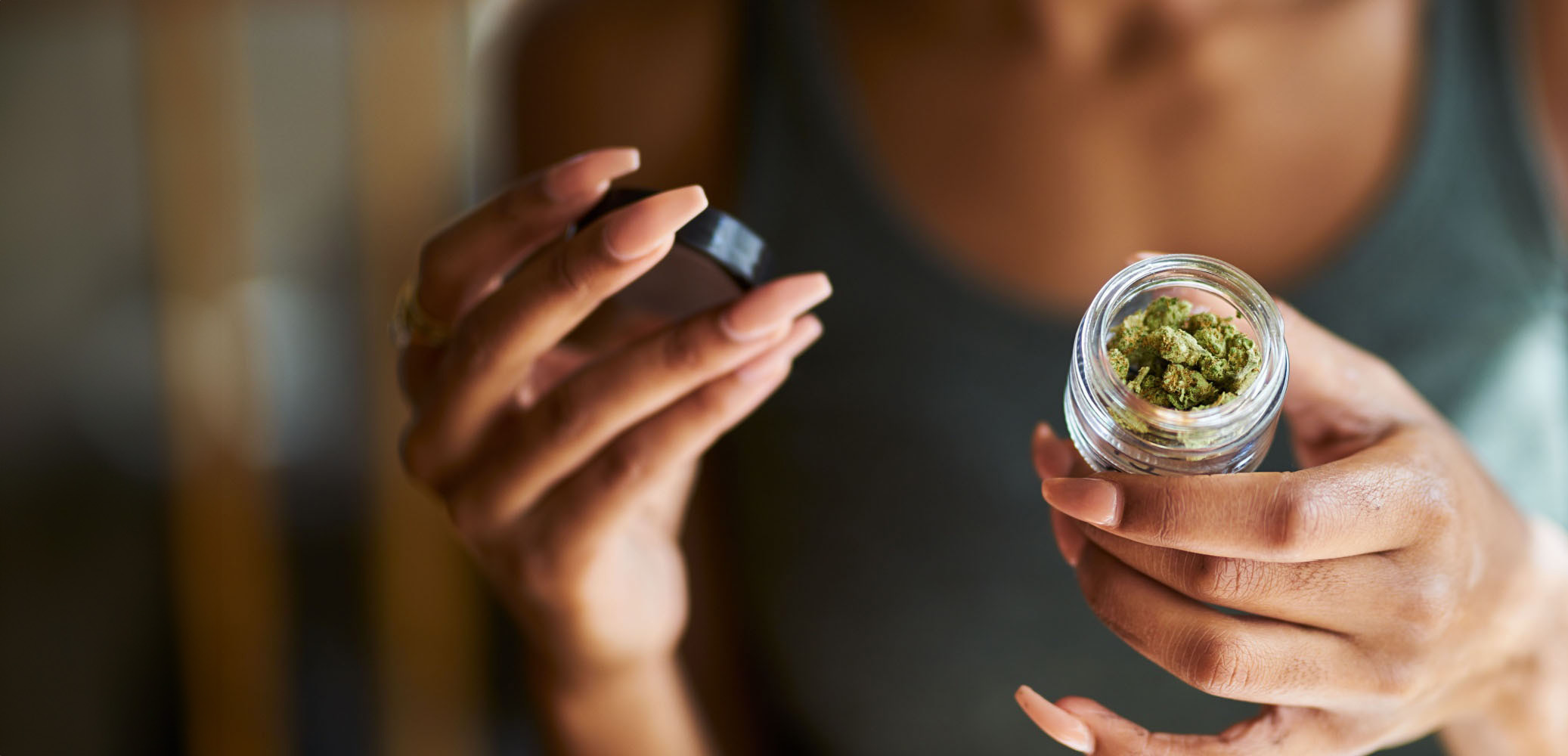 A person with long nails holds an open glass jar of cannabis buds—fresh from a cannabis delivery NYC & Long Island, NY—in one hand and the lid in the other. The blurred background highlights their sleeveless top and focus on quality product.