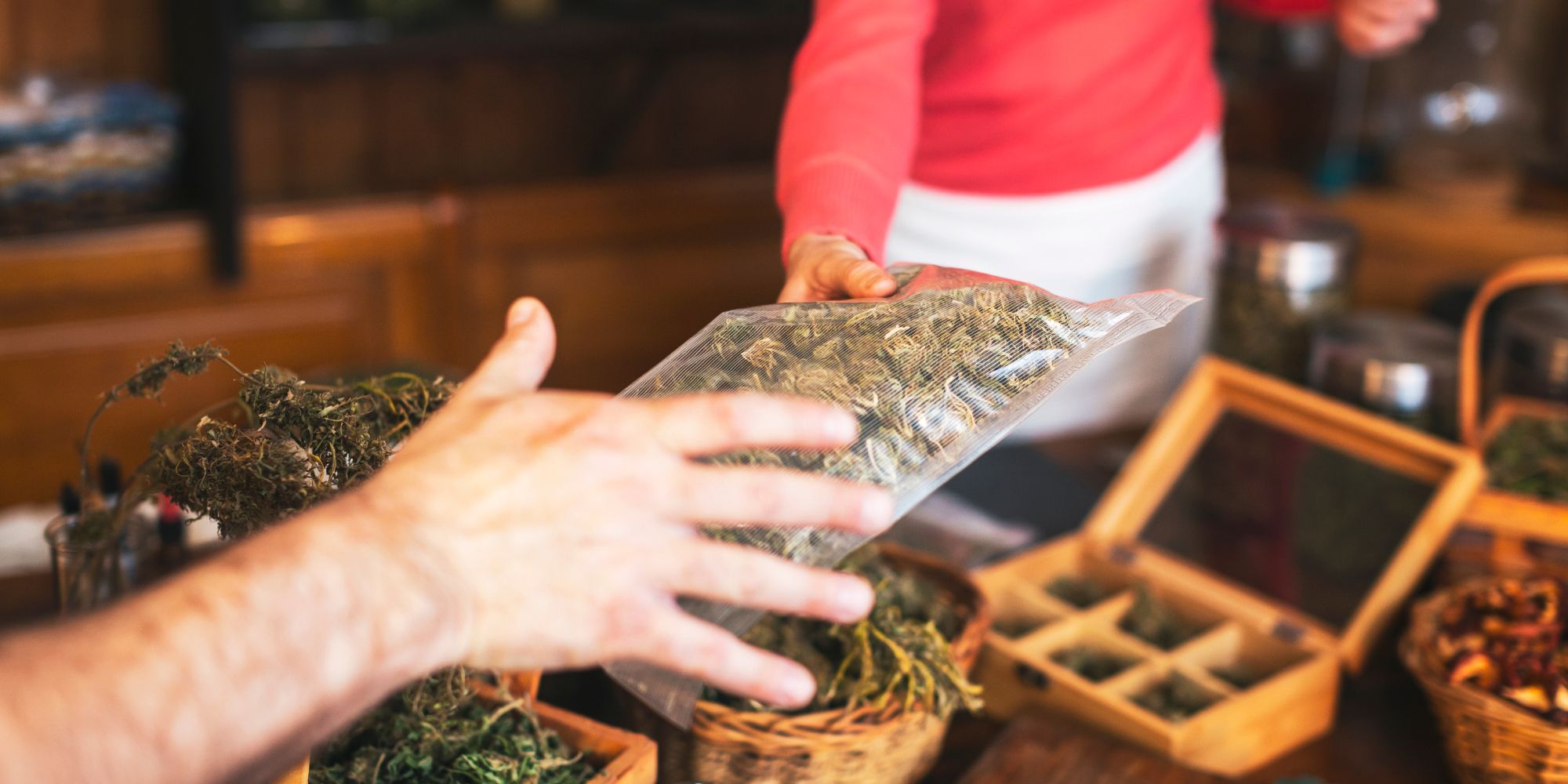 A person hands a clear plastic bag of dried cannabis to another across a counter in NY, with baskets of dried herbs and a wooden box on the table—capturing the essence of cannabis delivery NYC & Long Island style.