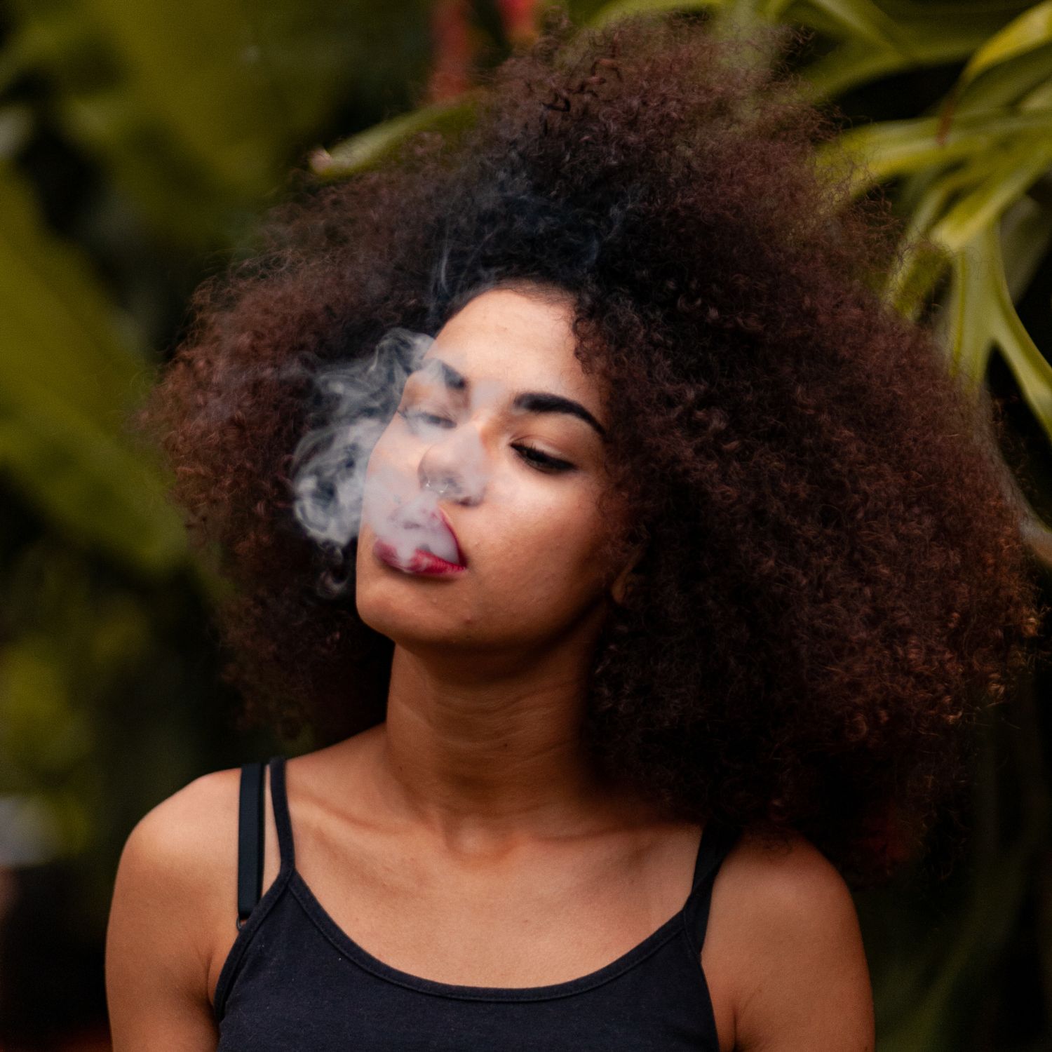 A person with curly hair, wearing a black tank top, exhales a cloud of smoke outdoors with green foliage in the background, enjoying their experience after using cannabis delivery NYC & Long Island, NY.