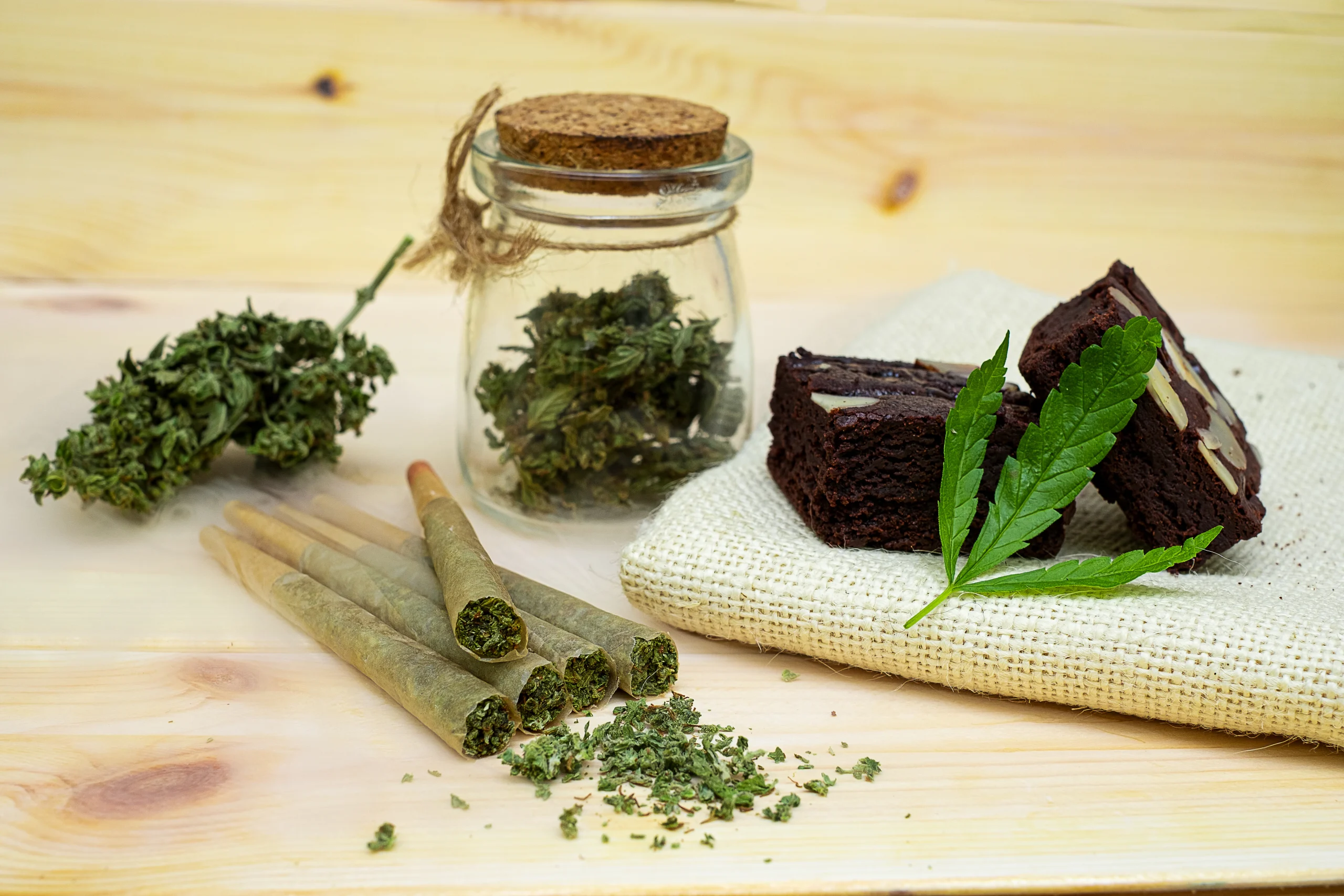 A jar of dried cannabis, marijuana buds, rolled joints, two brownies, and a cannabis leaf are arranged on a wooden surface with a beige cloth&mdash;perfect for those seeking cannabis delivery NYC & Long Island.