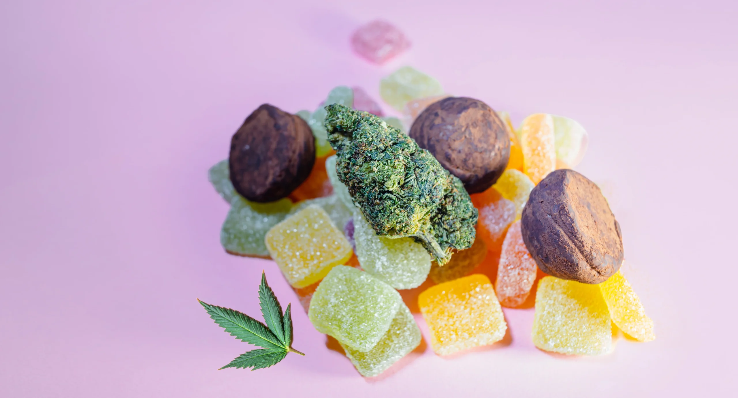 Assorted colorful gummy candies and chocolate truffles are arranged on a pink surface, topped with a cannabis bud and leaf—perfect treats available through cannabis delivery NYC & Long Island, NY.
