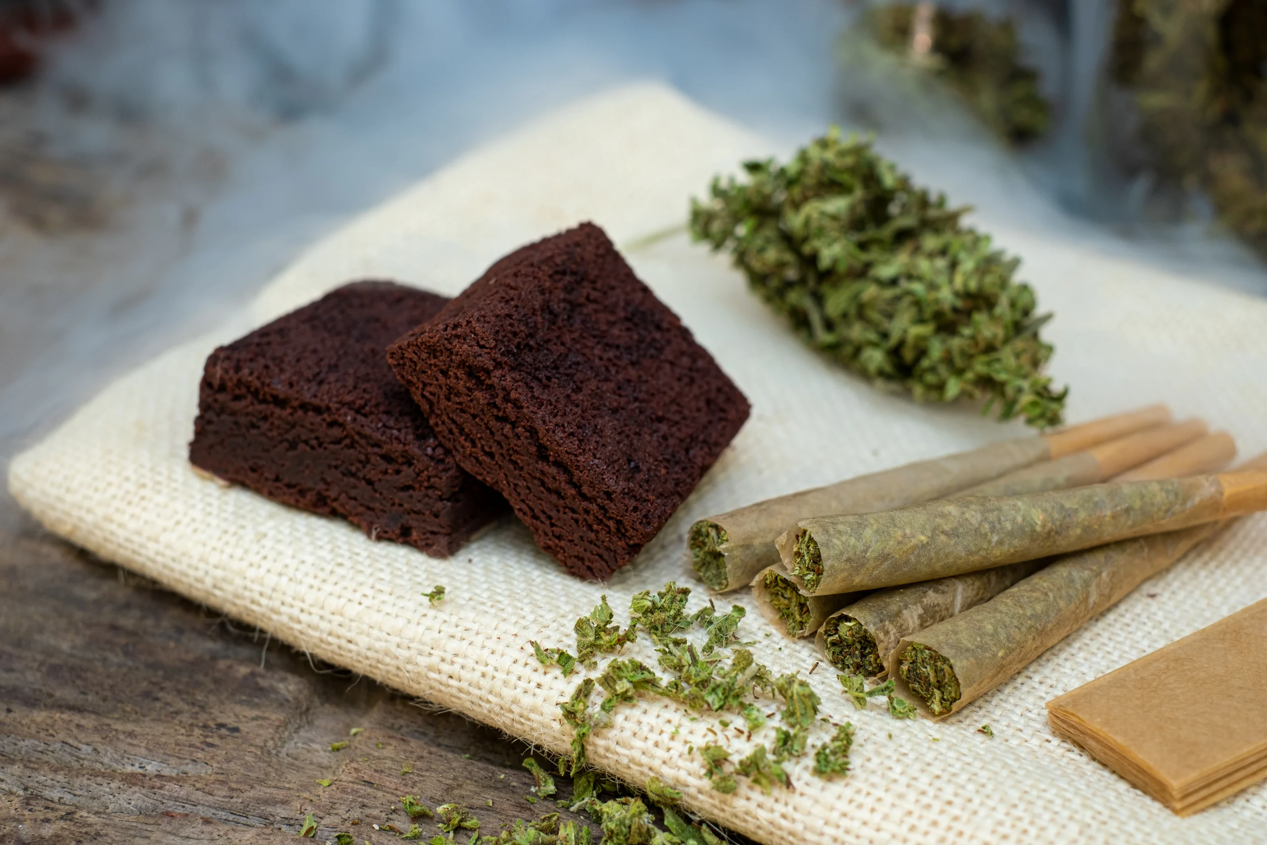 Two chocolate brownies on a textured cloth next to cannabis buds and three rolled joints, with some loose cannabis scattered nearby—perfect for relaxing while enjoying premium cannabis delivery NYC & Long Island.
