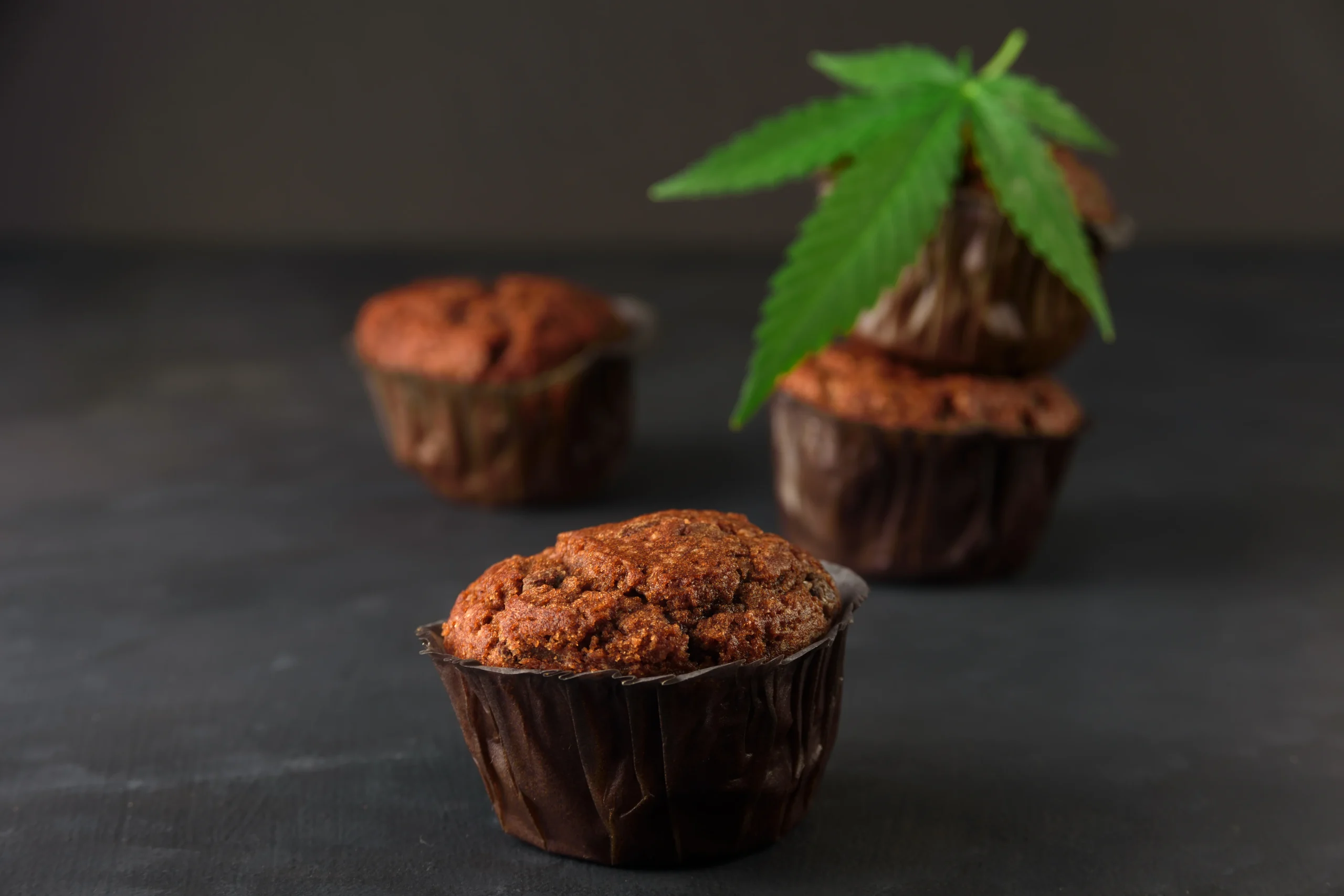 Chocolate muffins in dark wrappers are arranged on a moody surface, with one muffin topped by a green cannabis leaf&mdash;hinting at cannabis infusion. Perfect for those seeking cannabis delivery NYC & Long Island, NY. The background remains blurred and dark.