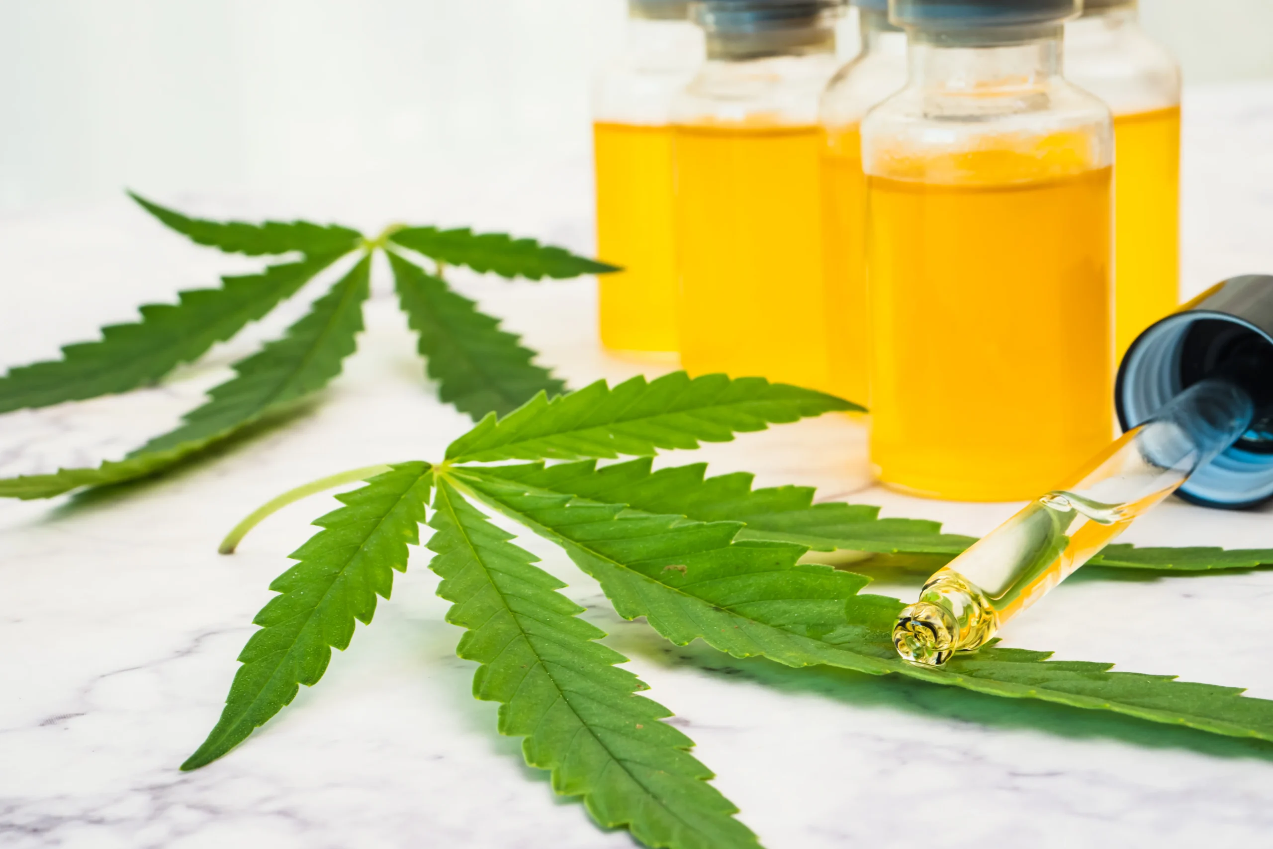Two cannabis leaves rest on a white marble surface near glass bottles filled with yellow liquid—a nod to premium cannabis delivery NYC & Long Island offers. A dropper, removed from one bottle, holds a visible drop of the golden extract at its tip.