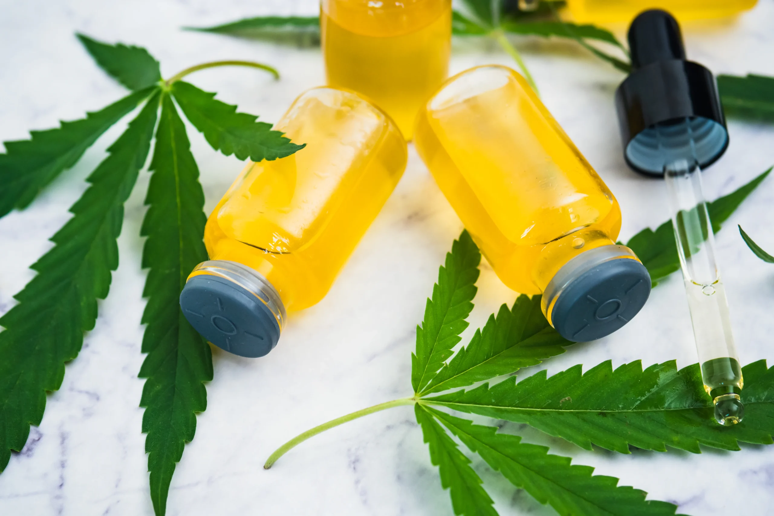 Small glass bottles filled with yellow liquid, a dropper, and green cannabis leaves are arranged on a white marble surface—perfect for showcasing premium products available through cannabis delivery NYC & Long Island, NY.