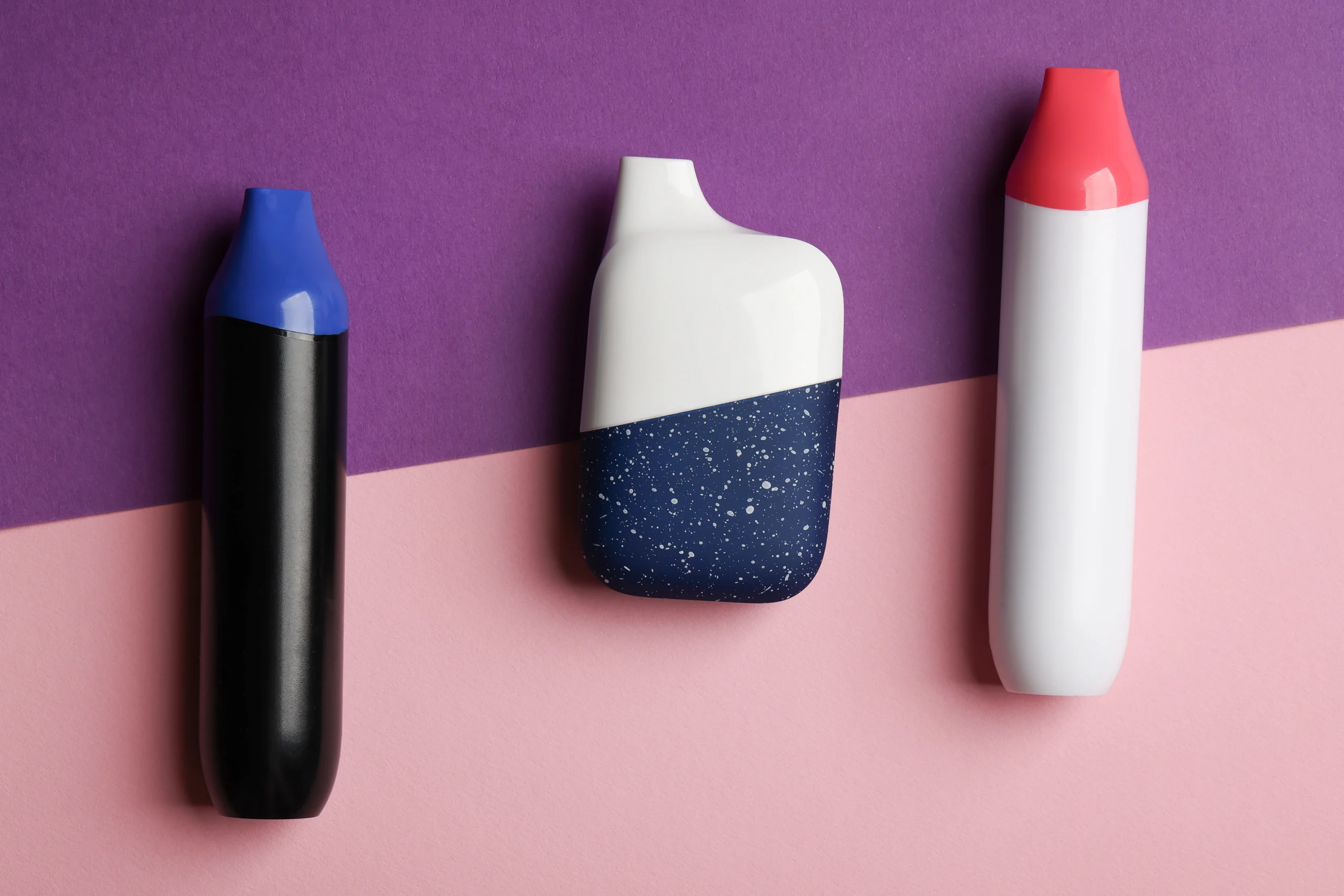 Three colorful vape pens are arranged vertically on a background split diagonally into purple and pink. Perfect for cannabis delivery NYC & Long Island, NY, the devices are black with blue, navy with white speckles, and white with a coral red tip.