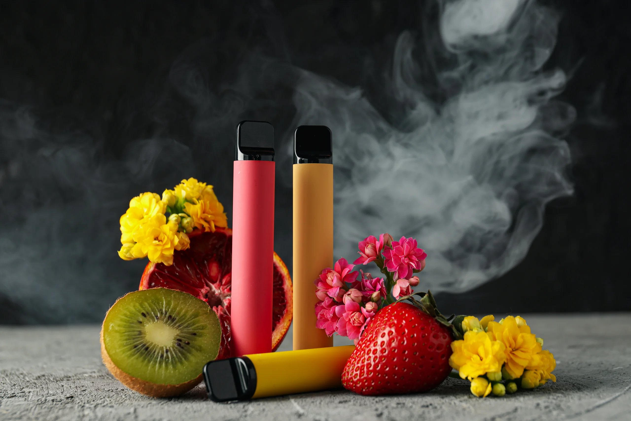 Three colorful vape pens stand upright among sliced kiwi, half a grapefruit, a whole strawberry, and bright flowers, with swirling smoke in the background—NY cannabis delivery NYC & Long Island brings these vibrant flavors to your door.