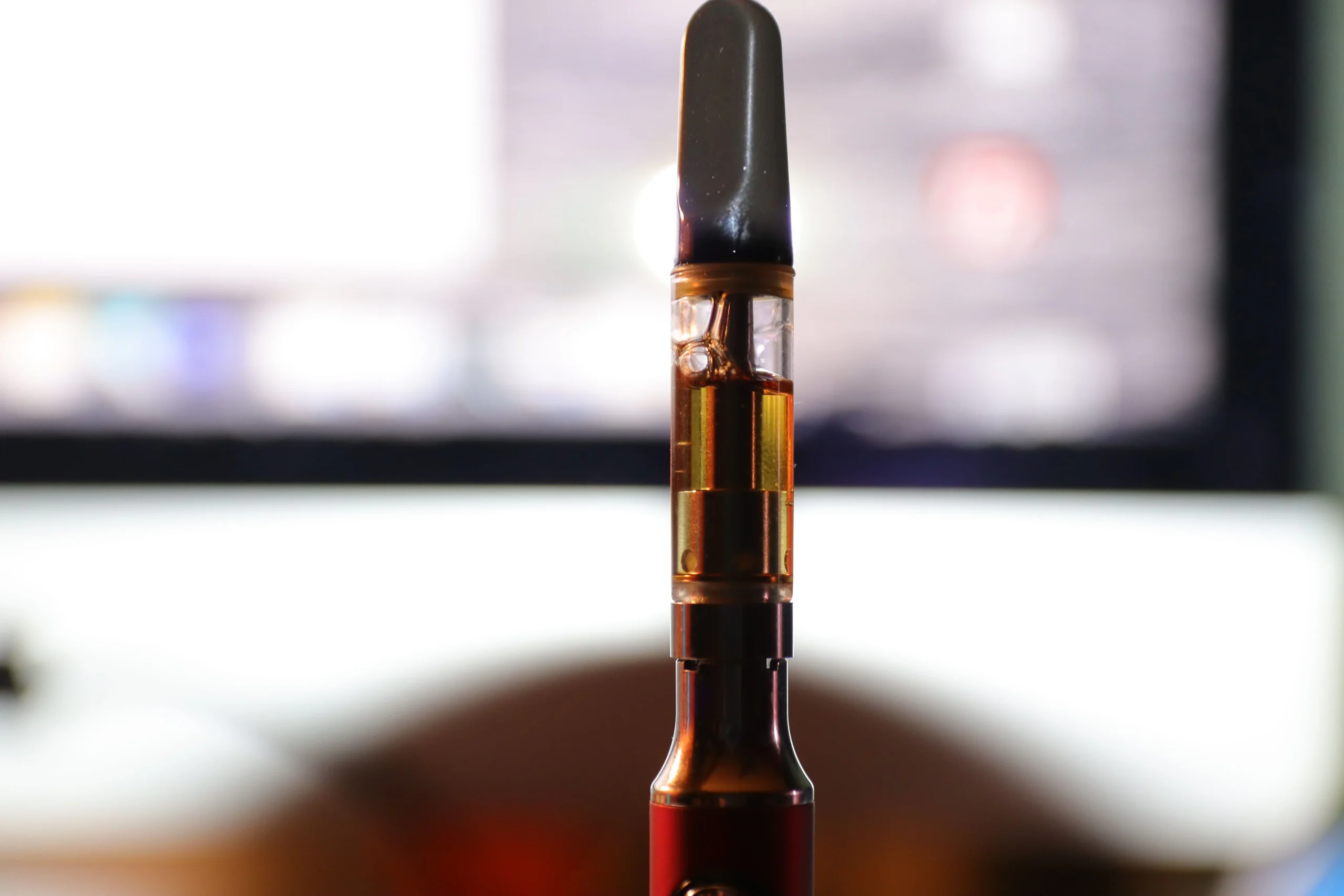 A close-up of a vape pen cartridge filled with amber liquid, set against the blurred background of an illuminated computer screen, highlights the convenience of cannabis delivery NYC & Long Island, NY.
