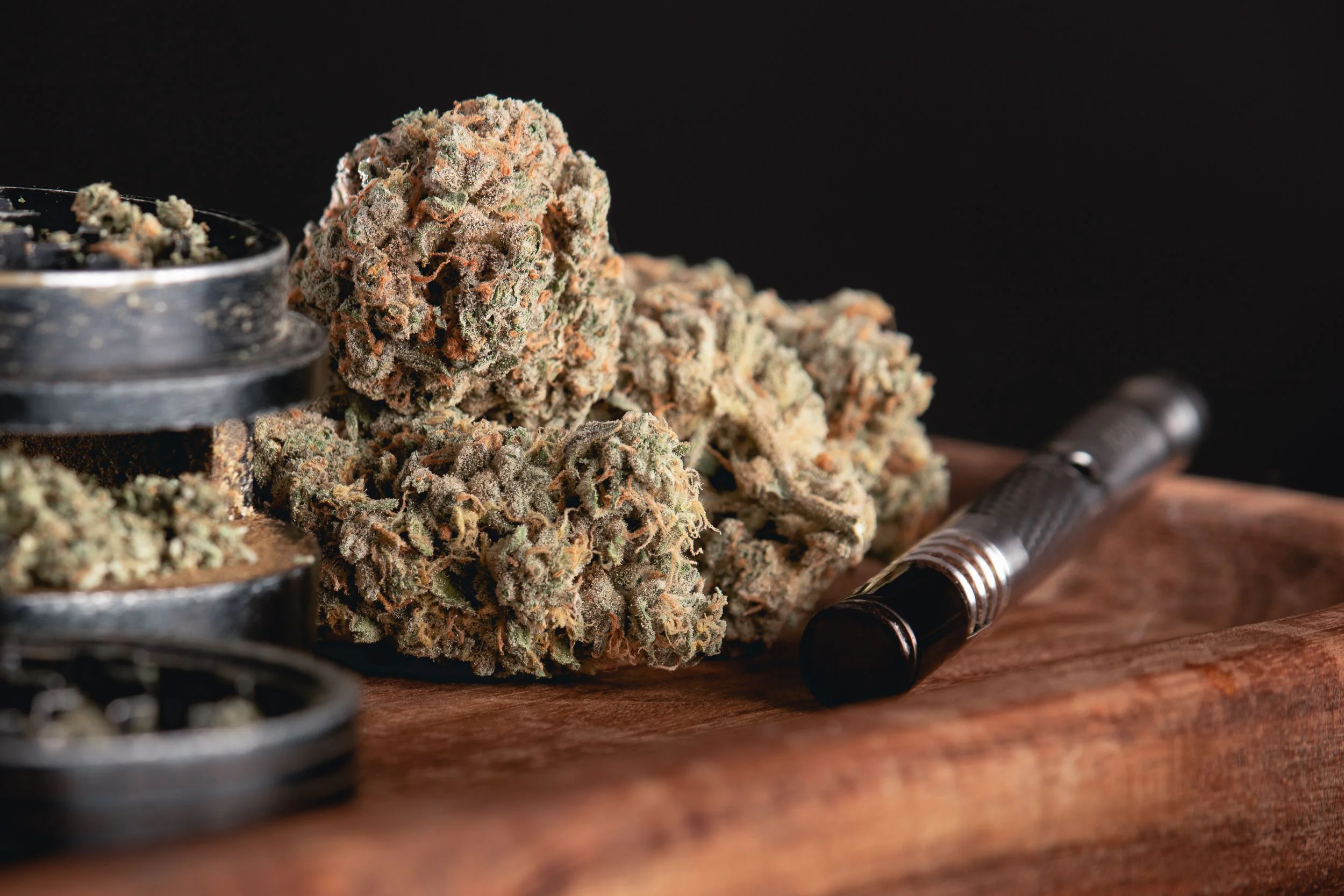 A close-up of dried cannabis buds on a wooden tray next to a metal vape pen and black grinder, with a dark background&mdash;perfect for those exploring NY cannabis delivery in NYC & Long Island.