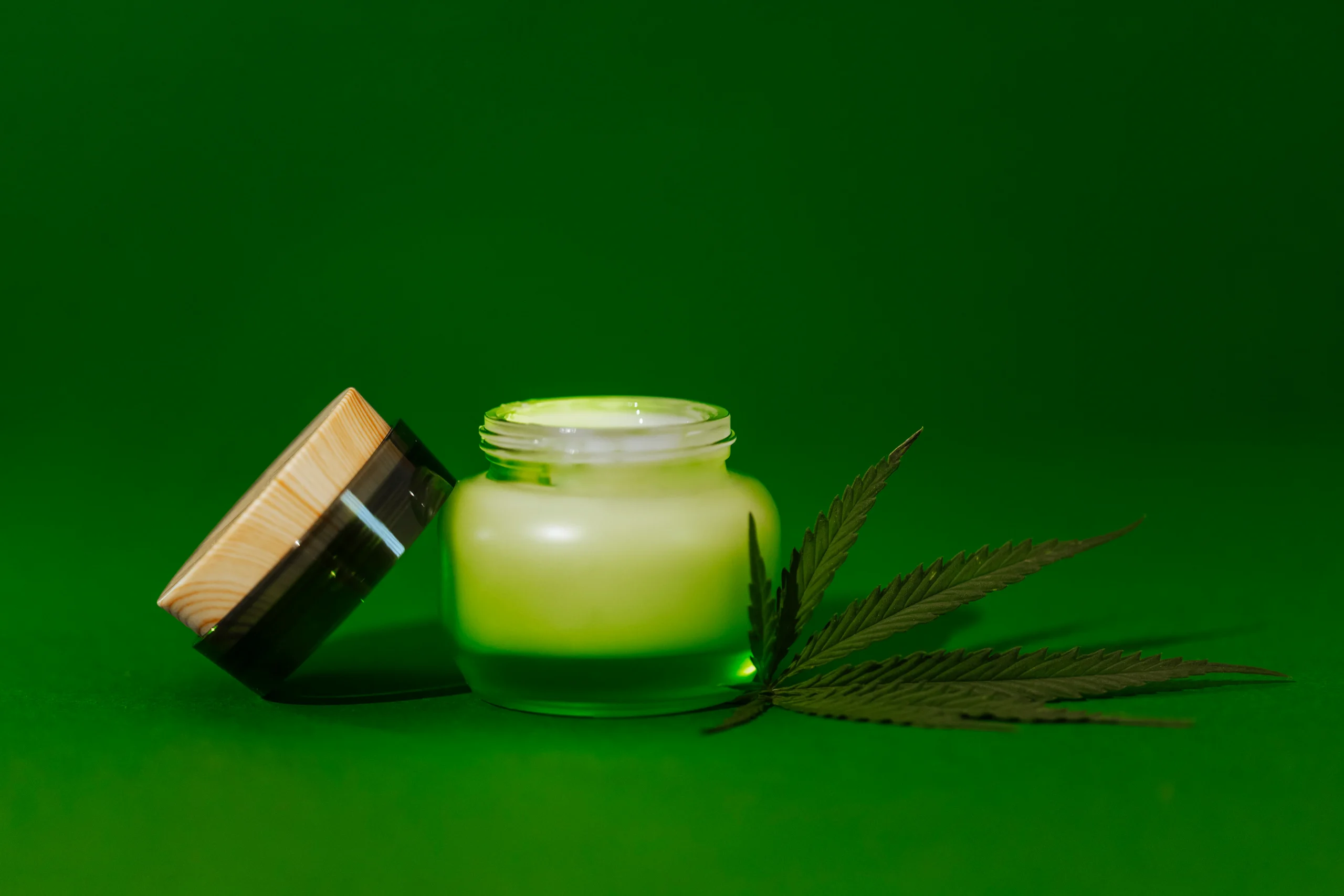 A jar of green cream with a wooden lid sits open on a green surface. Next to the jar is a cannabis leaf, highlighting premium cannabis delivery NYC & Long Island, all set against a matching green background.