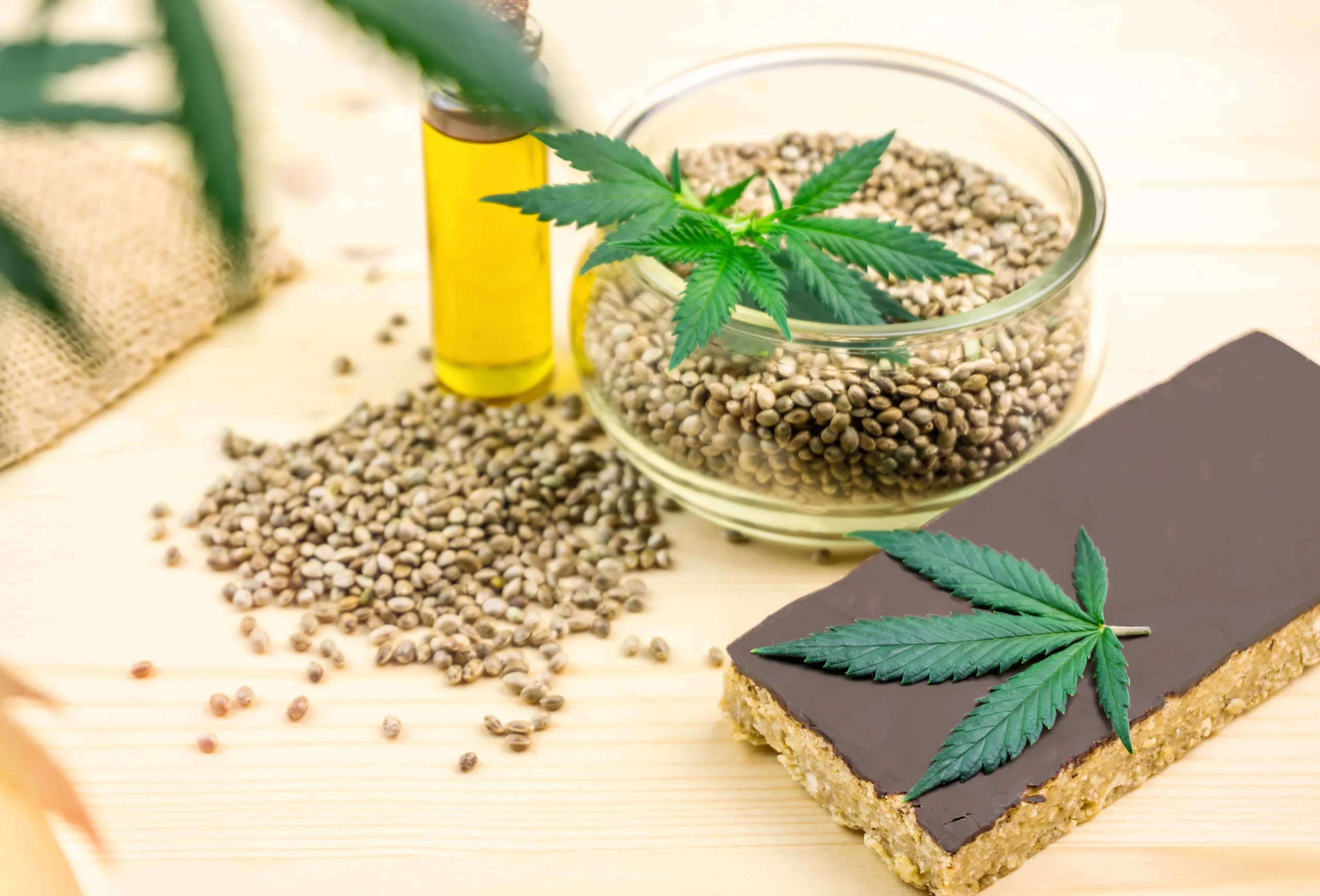 A glass bowl of hemp seeds, a small bottle of oil, and a chocolate-covered snack bar are arranged on a wooden surface with fresh cannabis leaves&mdash;a scene inspired by premium cannabis delivery NYC & Long Island, NY. Seeds are scattered nearby.
