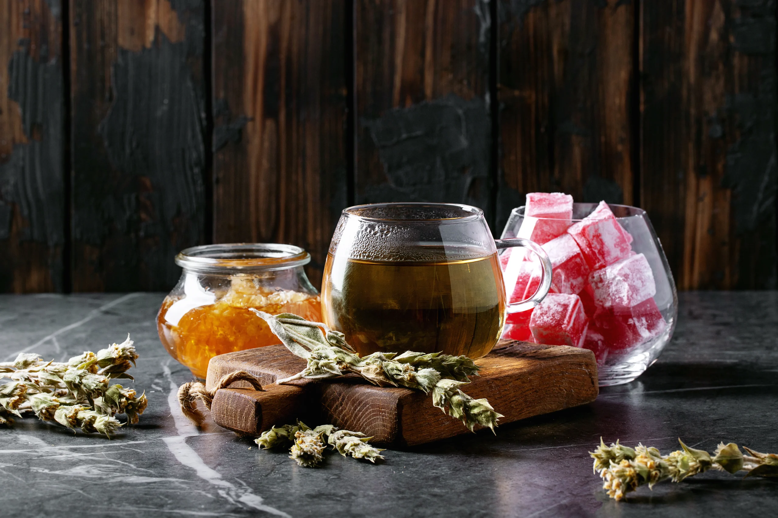 A glass mug of herbal tea, a jar of honey, dried herbs, and a bowl of pink Turkish delight cubes are arranged on dark marble—evoking the comfort and relaxation found with NY cannabis delivery in NYC & Long Island.