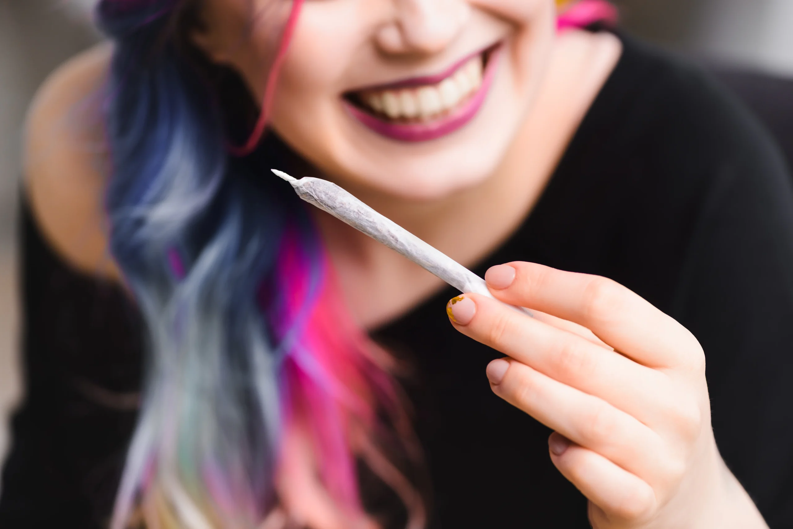 A smiling person with colorful hair holds a rolled joint in their hand—cannabis delivery NYC & Long Island bringing good vibes, their face slightly out of focus and the joint in sharp view.