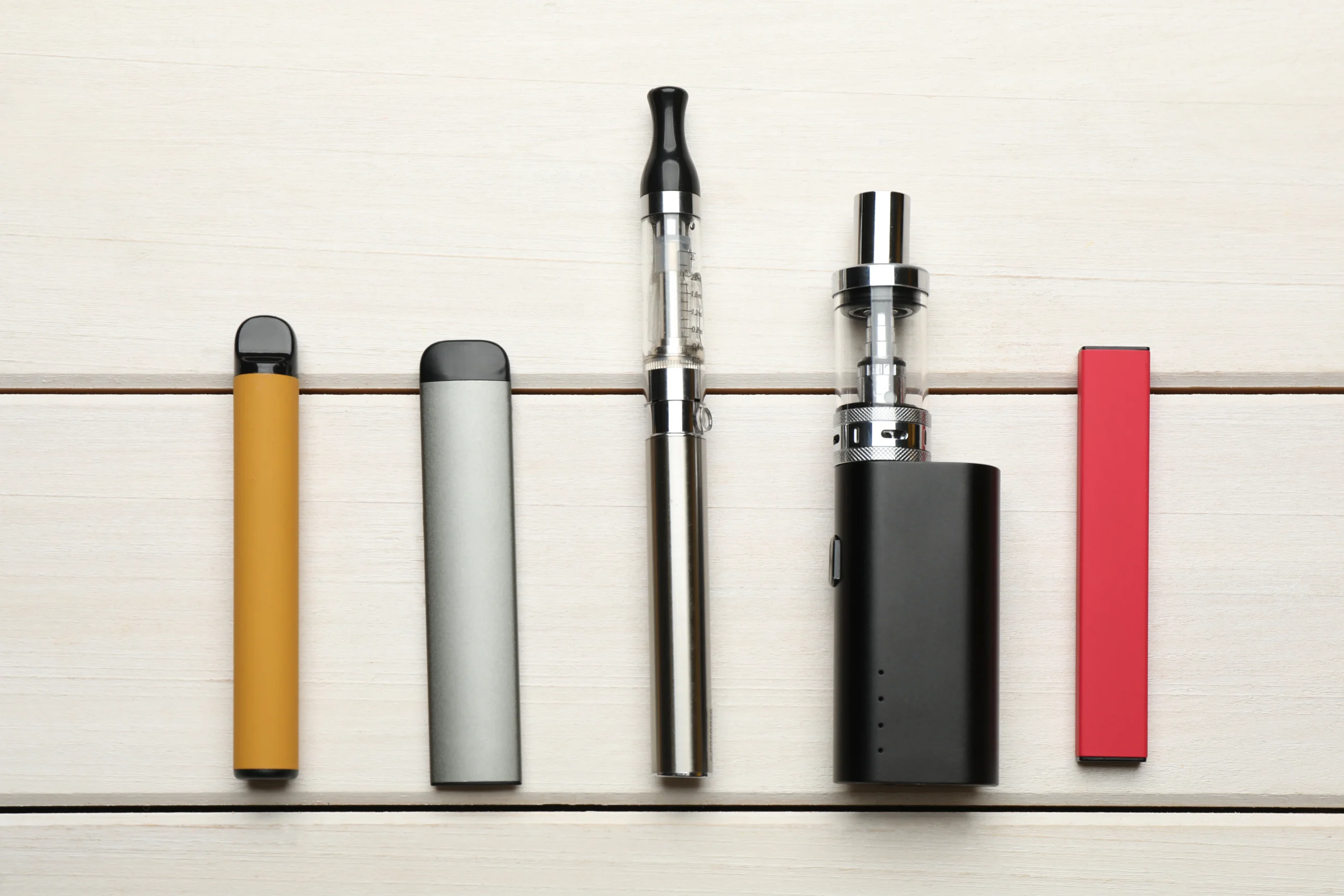 Five different vaping devices in various shapes and colors (yellow, gray, silver, black, and red) are arranged in a row on a light wooden surface&mdash;perfect for those seeking cannabis delivery NYC & Long Island options.