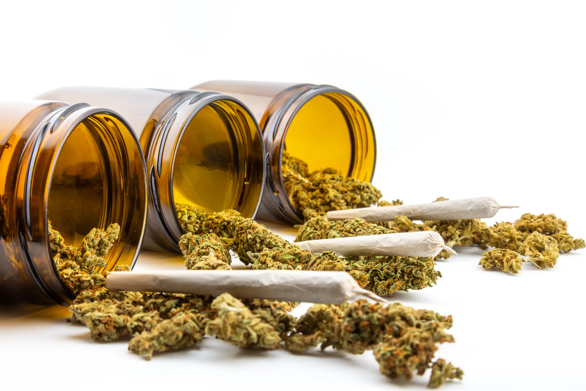 Three amber glass jars tipped over on their sides spill dried cannabis buds, with three rolled joints placed among the scattered buds, all set against a white background.