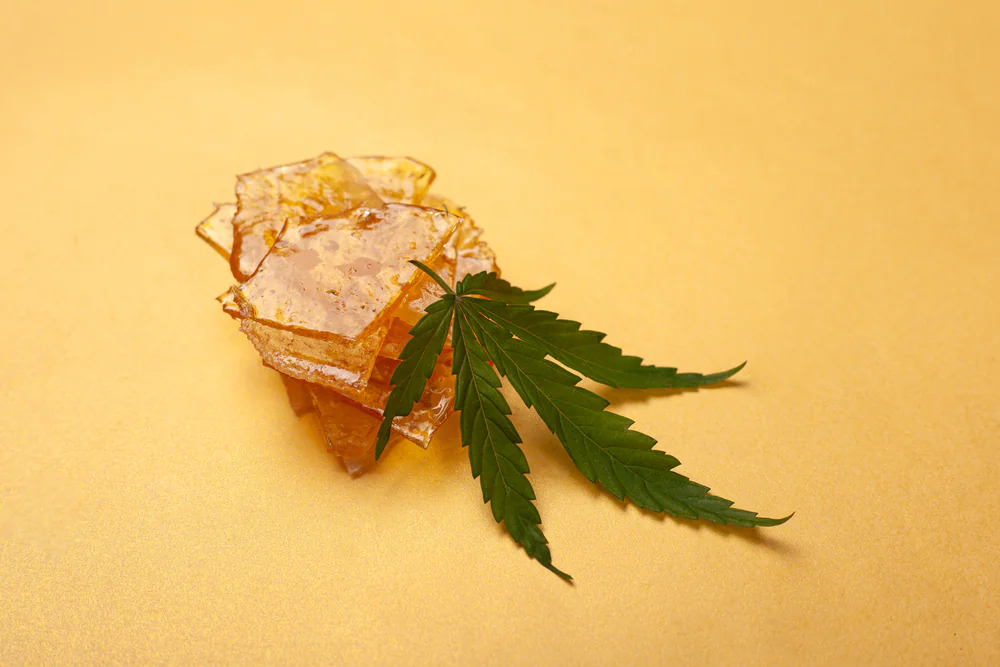 A stack of amber-colored cannabis concentrate shards topped with a green cannabis leaf, placed on a plain light yellow background.