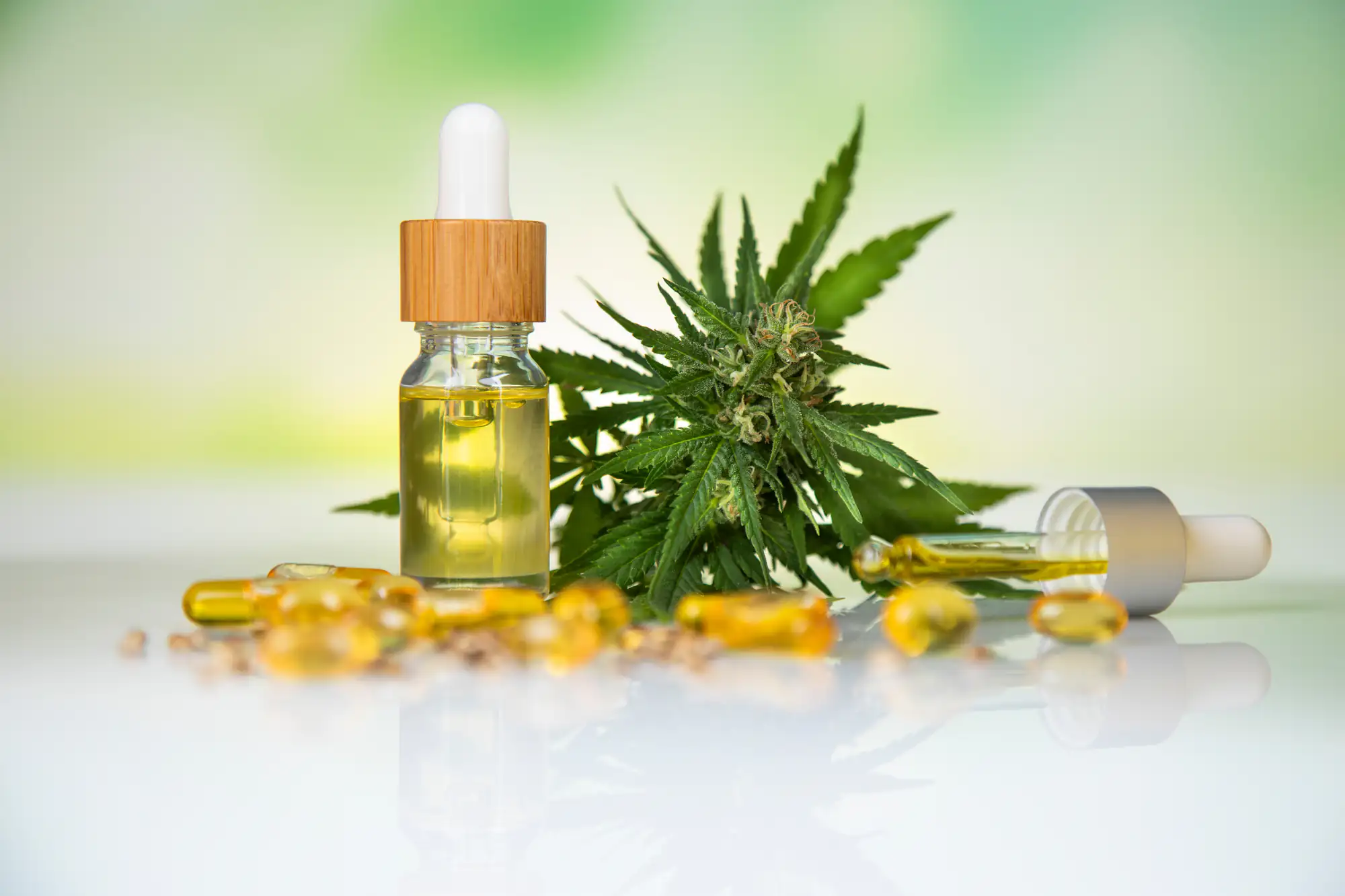 A small glass dropper bottle of oil, capsules, and a cannabis plant leaf and flower arranged on a white surface with a blurred green background.