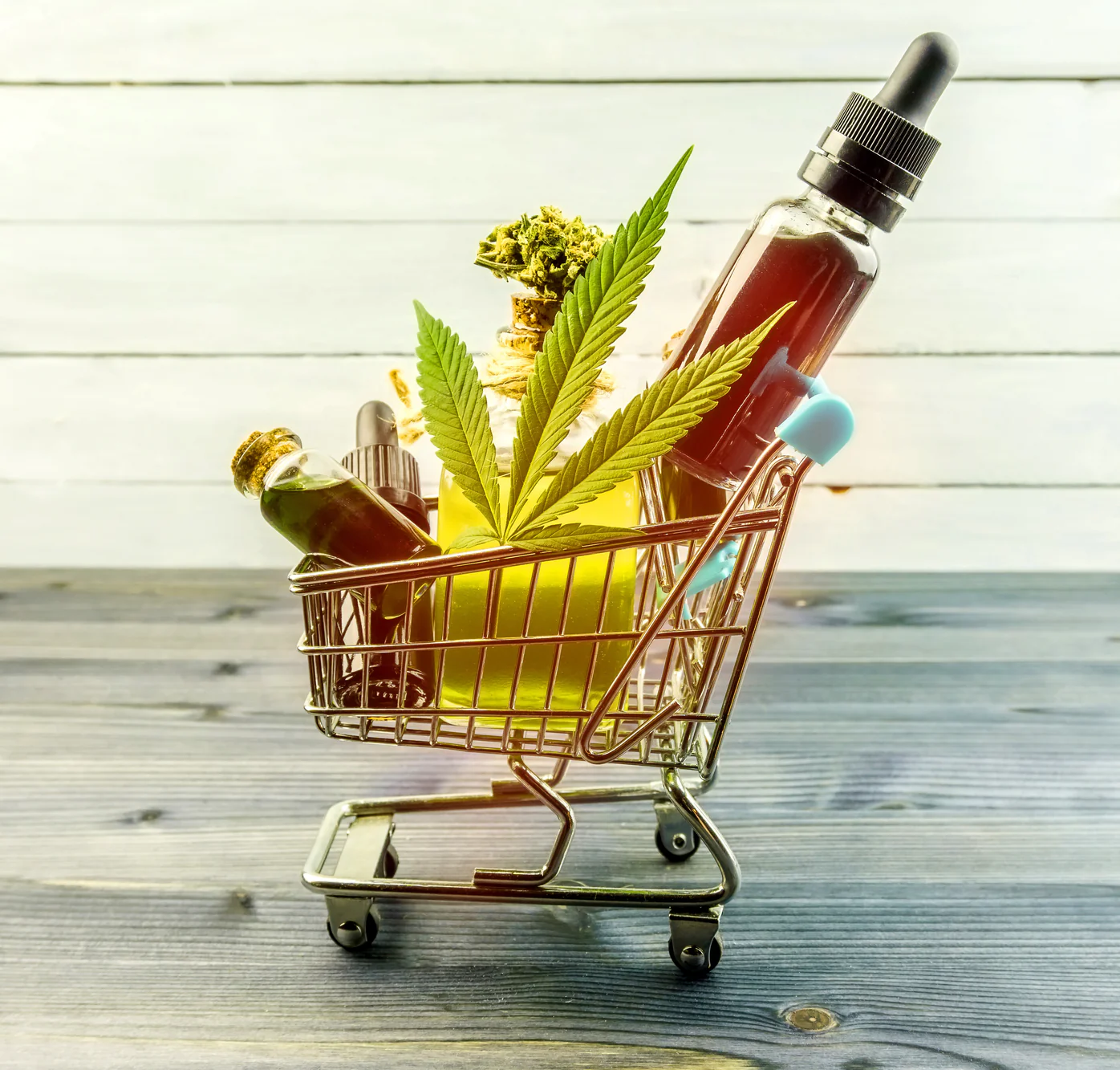A small shopping cart holds a cannabis leaf, a cannabis bud, glass bottles with liquid, and a dropper bottle, all placed on a wooden surface with a white wooden background.