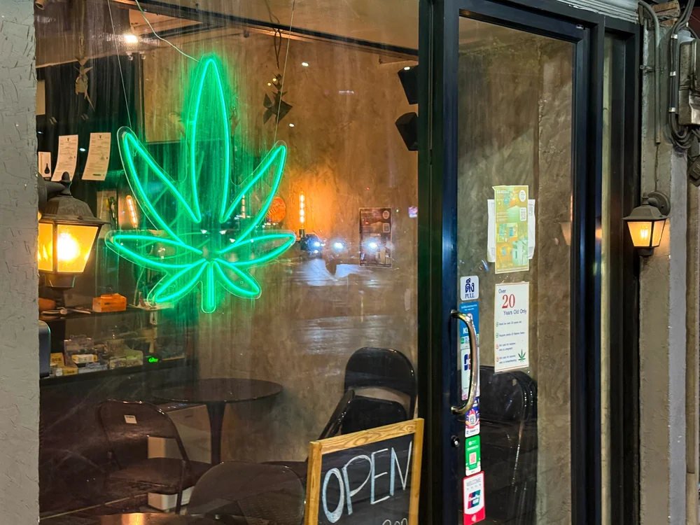 A storefront window with a bright green neon cannabis leaf sign, an "OPEN" sign on a chalkboard, and visible reflections of city lights at night. Tables and chairs are inside the shop.