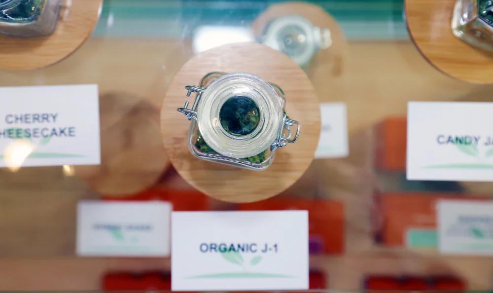 A glass jar filled with green plant material sits on a wooden coaster, labeled "ORGANIC J-1." Other jars and labels, including "CHERRY CHEESECAKE," are partially visible on either side.
