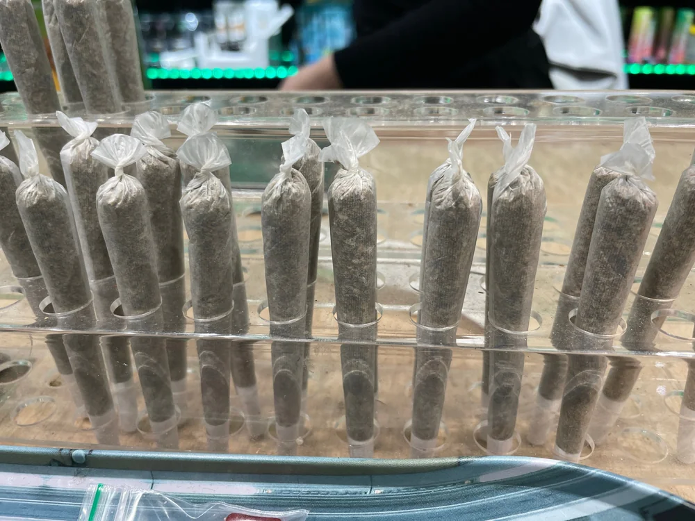 A row of pre-rolled joints stands upright in a clear plastic display case at a store counter, with jars and products visible in the background.
