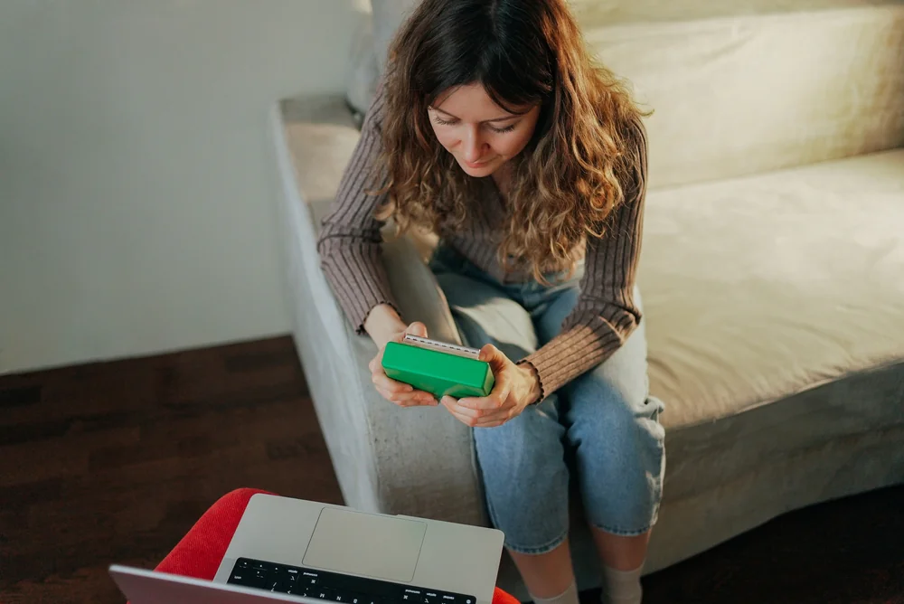 A woman with wavy hair sits on a beige couch, holding a green smartphone and looking at it, with an open laptop in front of her on a red table.