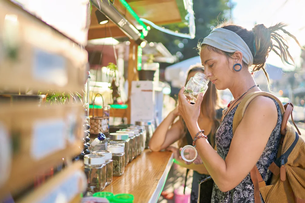 A person with a blue headband and backpack smells a jar at an outdoor market stall lined with jars. Sunlight and greenery are visible in the background.
