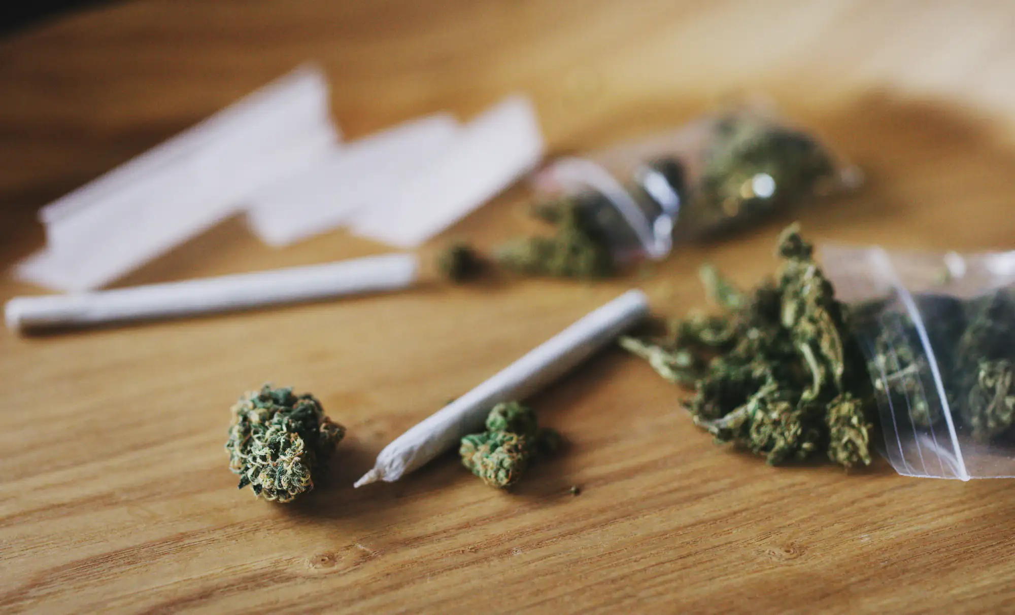 Loose marijuana buds, two rolled joints, empty rolling papers, and small plastic bags with cannabis are scattered on a wooden surface.