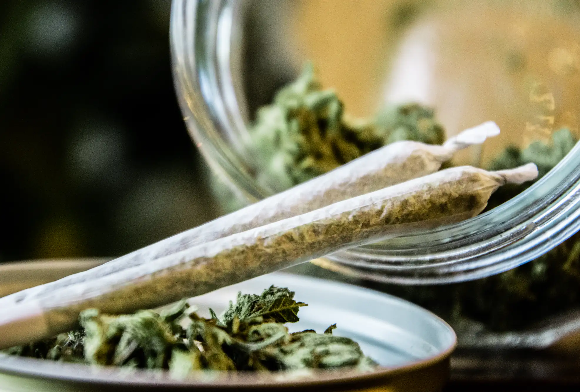 Two rolled cannabis joints rest on a metal lid next to loose cannabis buds spilling out of a glass jar. The image is focused on the joints and cannabis, with a blurred background.