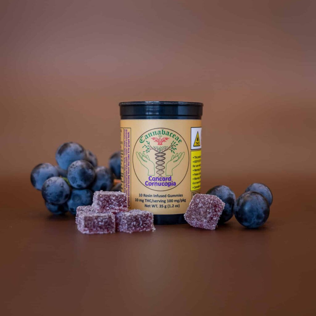 A container labeled “Concord Cornucopia” sits among clusters of grapes and purple sugar-coated gummies, all arranged on a brown surface with a plain brown background.