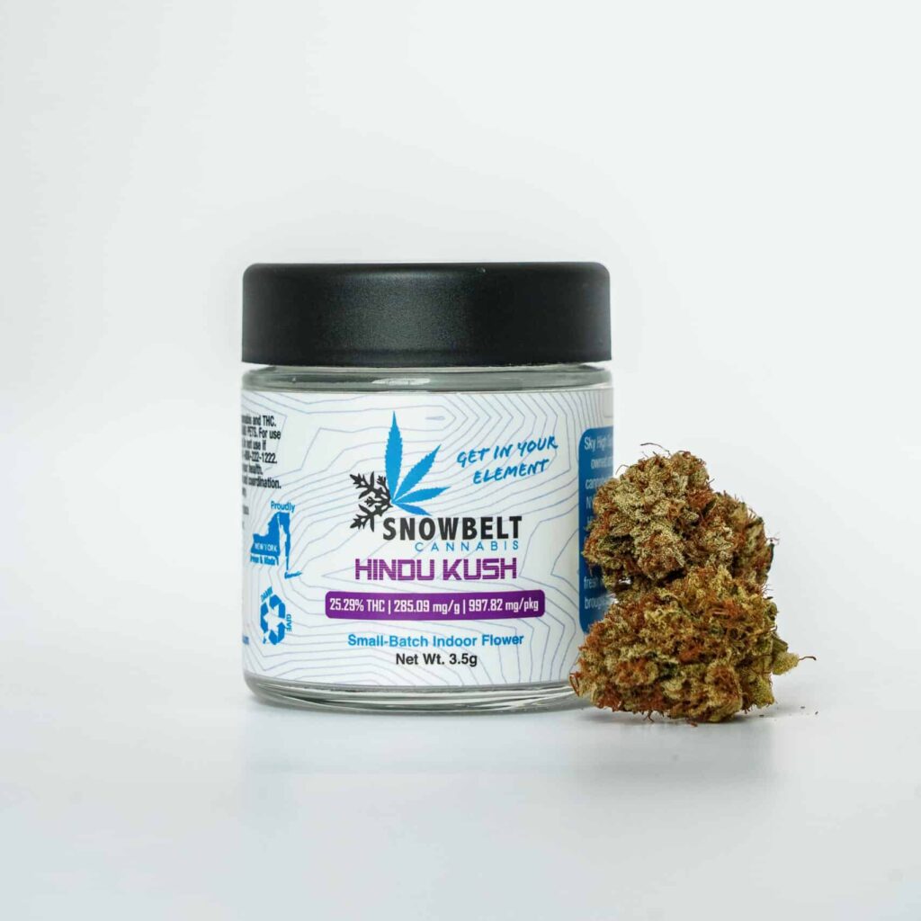 A glass jar labeled "Snowbelt Cannabis Hindu Kush" with product details sits next to a small pile of cannabis buds on a white background.