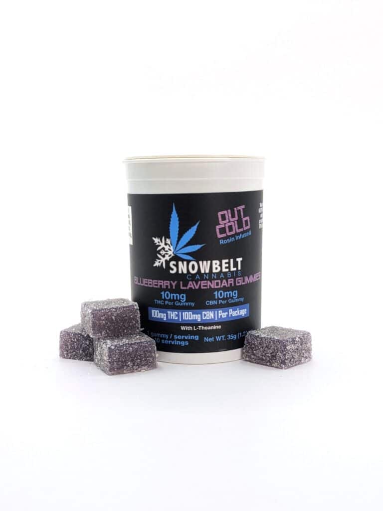 A white container labeled "Snowbelt Cannabis Blueberry Lavender Gummies" stands upright, with five purple sugar-coated gummy cubes arranged in front of it on a white background.