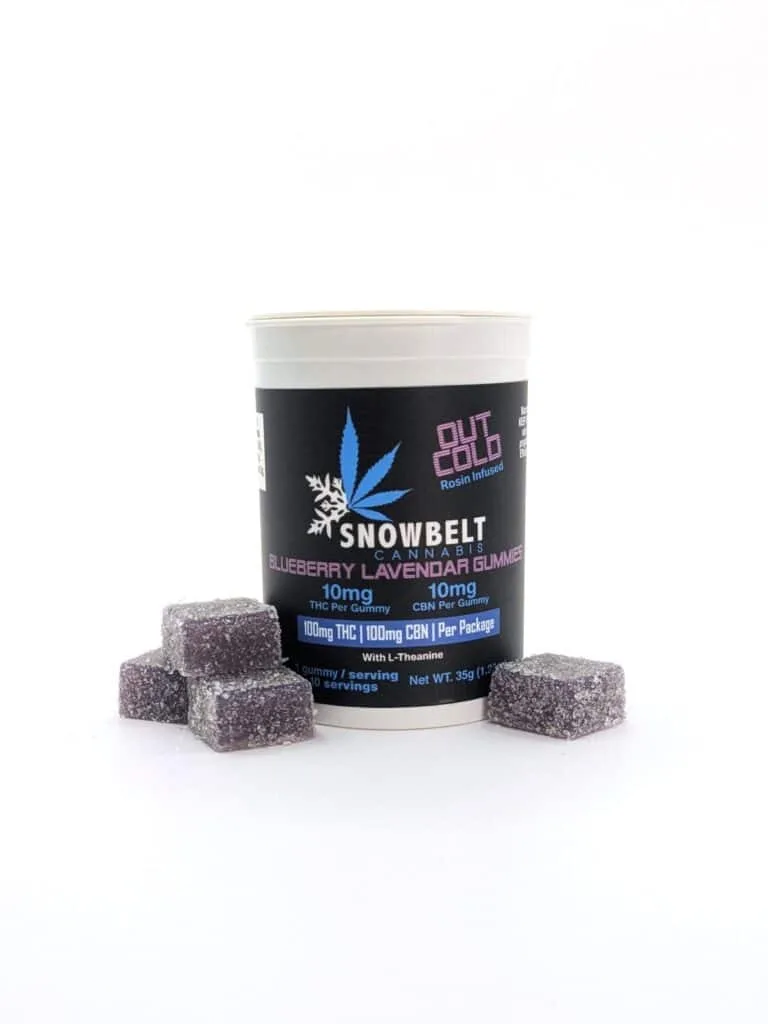 A white container labeled "Snowbelt Cannabis Blueberry Lavender Gummies" stands upright, with five purple sugar-coated gummy cubes arranged in front of it on a white background.