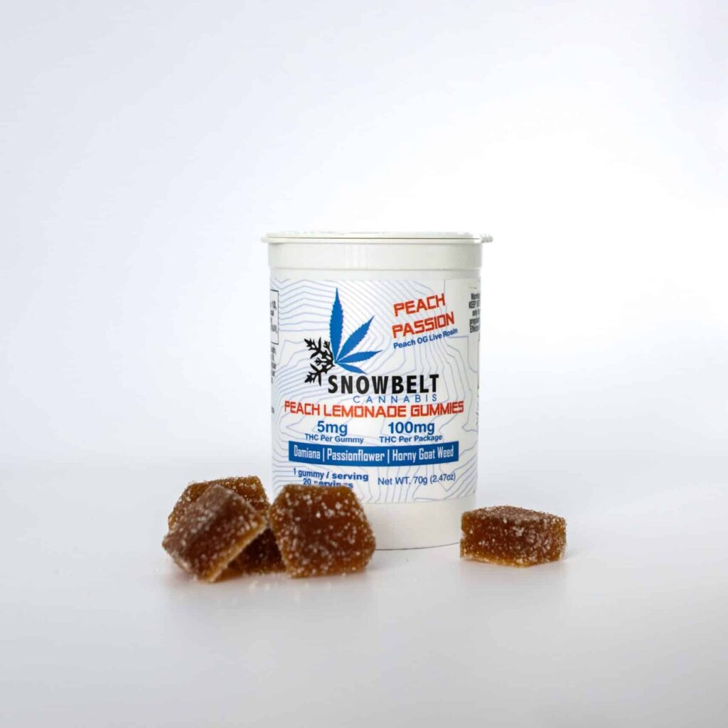 A white container labeled "Snowbelt Cannabis Peach Passion Peach Lemonade Gummies" stands upright, with several sugar-coated gummies arranged in front of it on a white background.