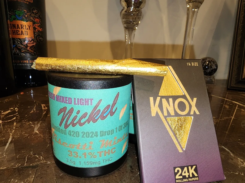 A gold-wrapped joint rests on a black jar labeled “Nickel” next to a black and gold box labeled “KNOX 24K rolling papers” on a marble surface, with bottles and glassware in the background.