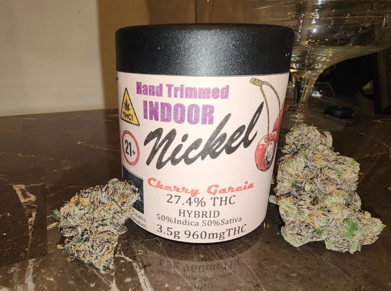 A black container labeled "Hand Trimmed Indoor Nickel Cherry Garcia" with 27.4% THC, Hybrid (50% Indica, 50% Sativa), 3.5g 960mg THC. Two cannabis buds are placed beside the container on a marble surface.