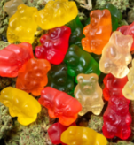 Colorful gummy bears in red, yellow, orange, green, and clear are scattered on a surface covered with cannabis buds.