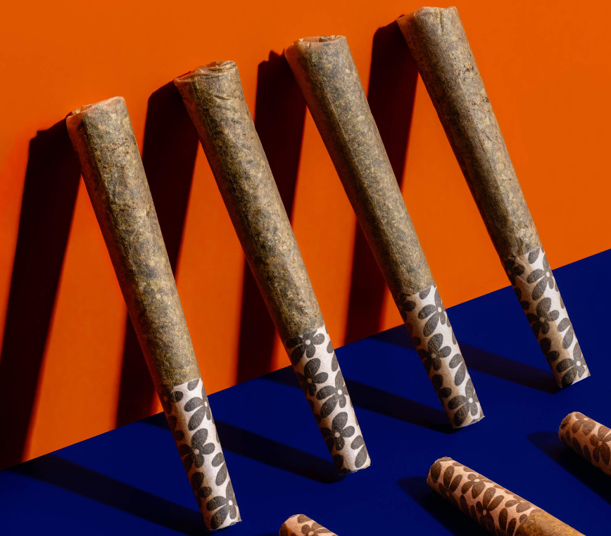 Four rolled joints with decorative patterned tips are arranged diagonally on a background divided into orange and blue sections. Shadows are cast on the surface, and parts of additional joints are visible at the bottom edge.