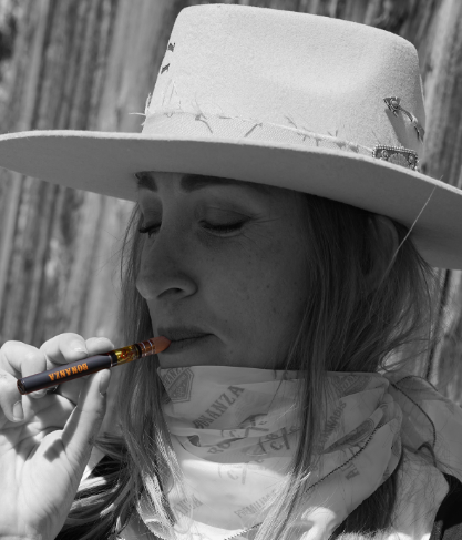 A person wearing a light wide-brimmed hat and a bandana around their neck holds a vape pen to their lips. The image is in black and white, except for the colored vape pen labeled "BURNNA.
