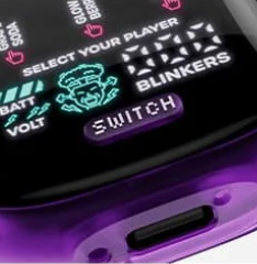 Close-up of an electronic device with a digital display showing the words "SELECT YOUR PLAYER," "BLINKERS," battery and voltage indicators, and a "SWITCH" button, all on a glossy purple and black surface.