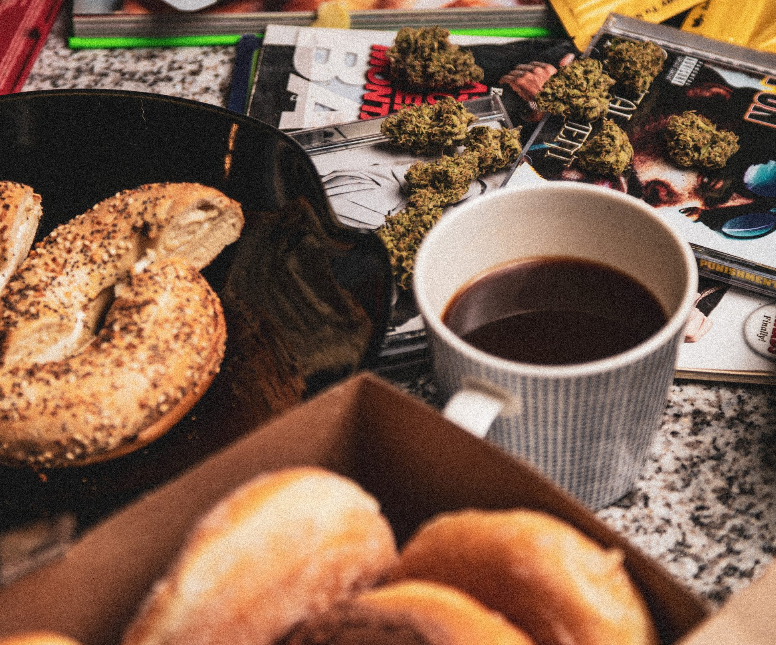 A table with a plate of bagels, a box of donuts, a mug of coffee, magazine covers, and several cannabis buds scattered on the surface.