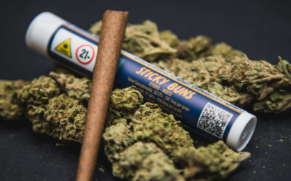 A labeled tube reading "Sticky Buns" lies among cannabis buds and a rolled blunt on a dark surface. The tube shows a 21+ warning and a QR code.