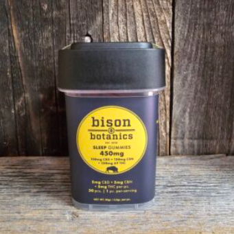 A container of Bison Botanics Sleep Gummies sits on a rustic wooden surface. The label is yellow with black text, stating the gummies contain 450mg CBD and CBN, and the container has a black lid.