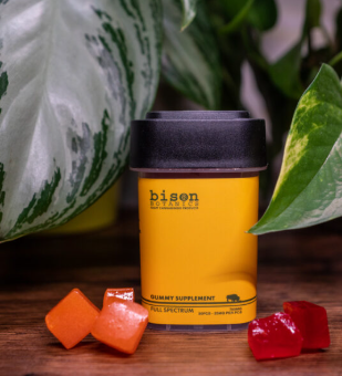 A yellow container labeled "Bison Botanics Gummy Supplement Full Spectrum" is surrounded by red and orange gummy cubes, with green leafy plants in the background.