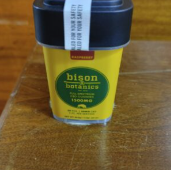 A yellow container of Bison Botanics Full Spectrum CBD Gummies, raspberry flavor, 1500mg, with a black lid and a white safety seal, placed on a wooden surface.