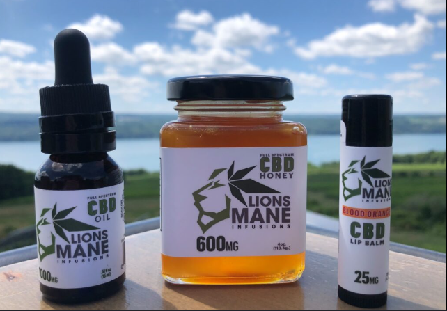 Three Lions Mane CBD products—oil (600mg), honey (600mg), and blood orange lip balm (25mg)—are displayed outdoors on a ledge with a scenic lake and greenery in the background under a partly cloudy sky.