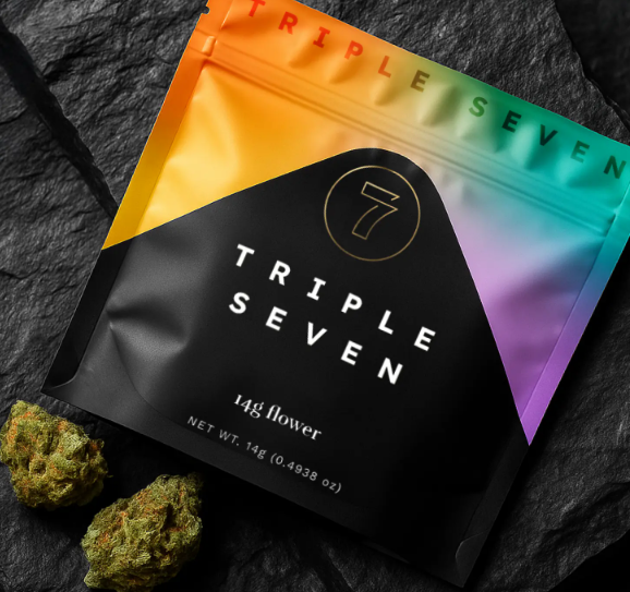 A colorful resealable pouch labeled "Triple Seven" with "14g flower" on the front, placed on a dark stone surface next to two cannabis buds.