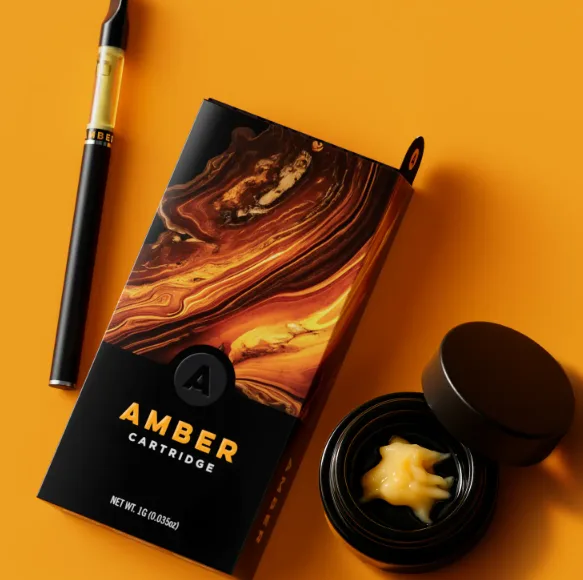 A vape pen, a black and amber swirl cartridge box labeled "Amber Cartridge," and a small open container with a creamy yellow substance are displayed on an orange surface.