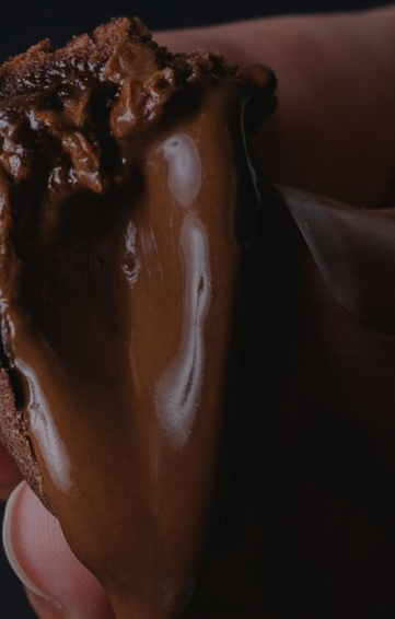 A close-up of a hand holding a piece of chocolate cake topped with smooth, glossy chocolate ganache, set against a dark background.