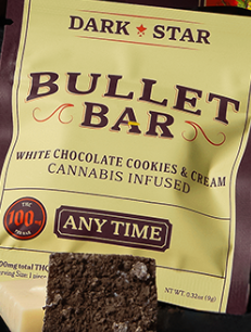 A package labeled "DARK STAR BULLET BAR" advertising a white chocolate cookies & cream cannabis-infused bar, with text highlighting "100+ mg THC" and "ANY TIME." A piece of the bar is in front of the package.